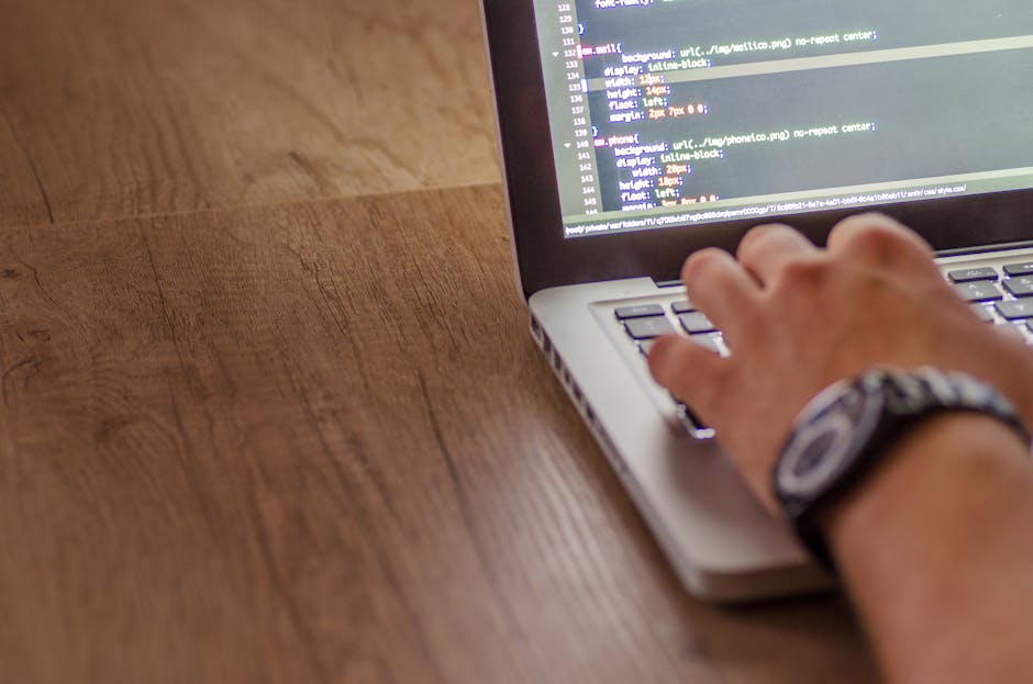 Close-up of a person's hand operating a silver laptop on a wooden desk with visible grain and texture. The screen displays lines of code with various colored syntax highlighting. The person wears a da