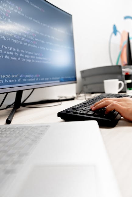Close-up of a person's hand operating a silver laptop on a wooden desk with visible grain and texture. The screen displays lines of code with various colored syntax highlighting. The person wears a da